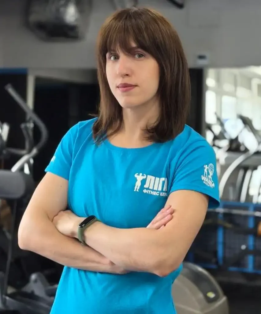 Woman with brown hair in a blue gym shirt stands with arms crossed in a fitness center, looking at the camera.