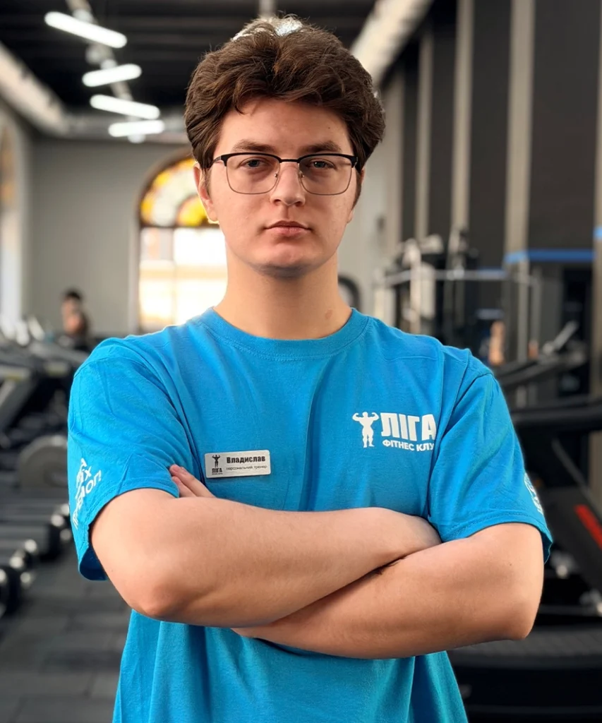Young man with short brown hair and glasses stands with arms crossed in a gym, wearing a blue shirt and a name badge.
