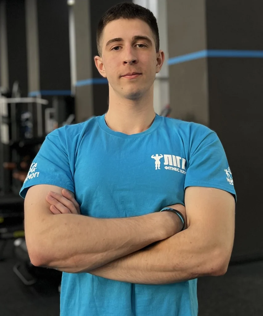 Young man with short dark hair stands in a gym with arms crossed, wearing a blue fitness shirt.