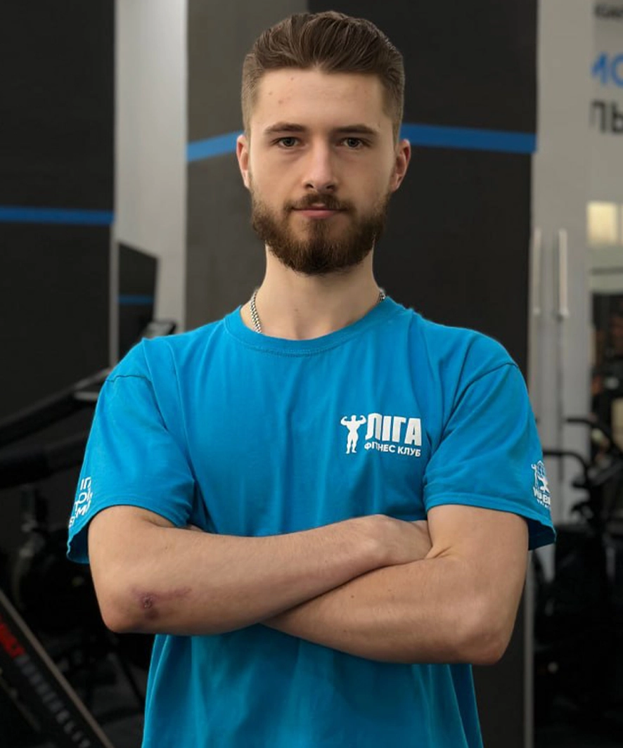 Young man with a beard standing in a gym with arms crossed, wearing a blue fitness club t-shirt.